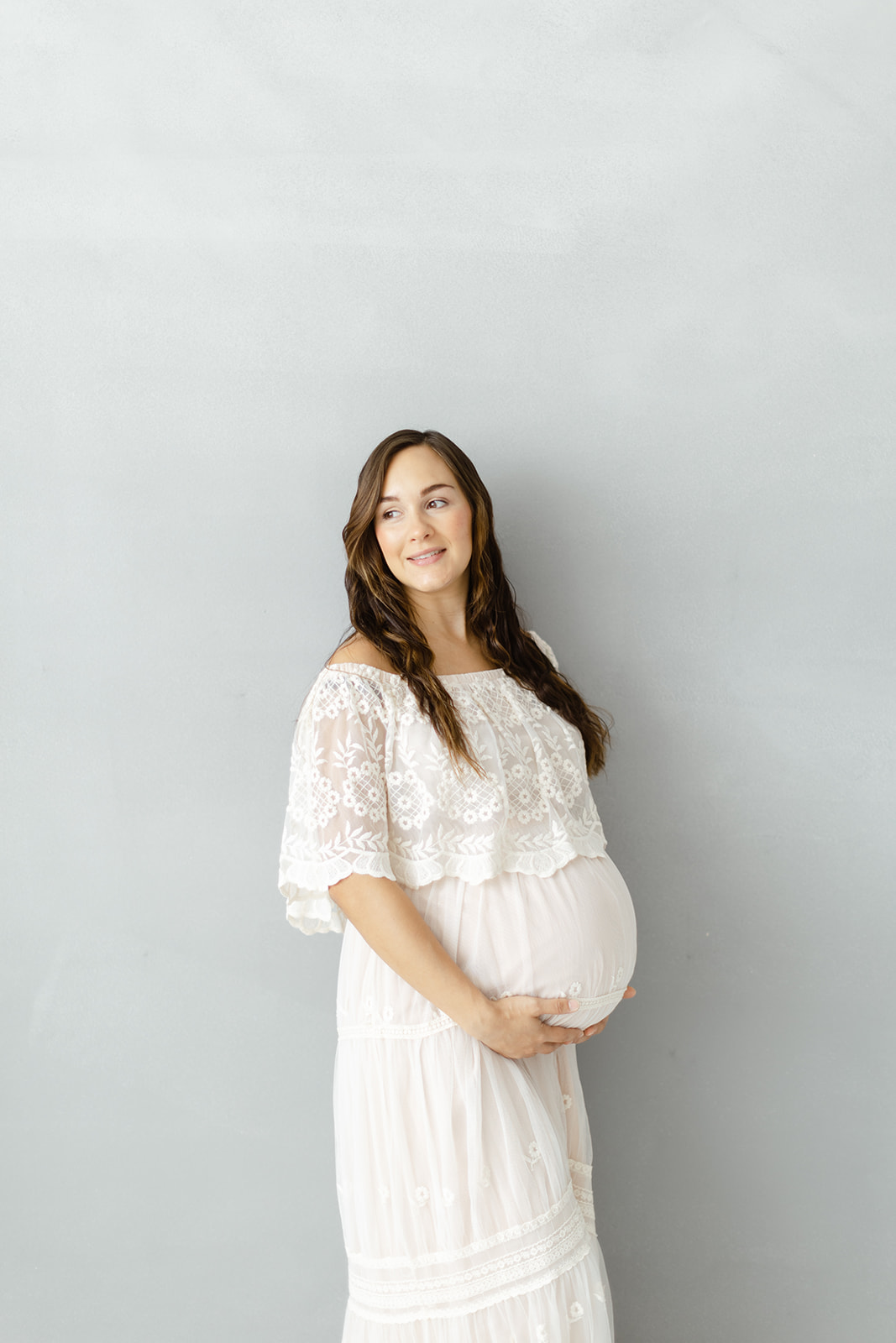 A mom to be holds the bump while standing in a studio in a white maternity gown