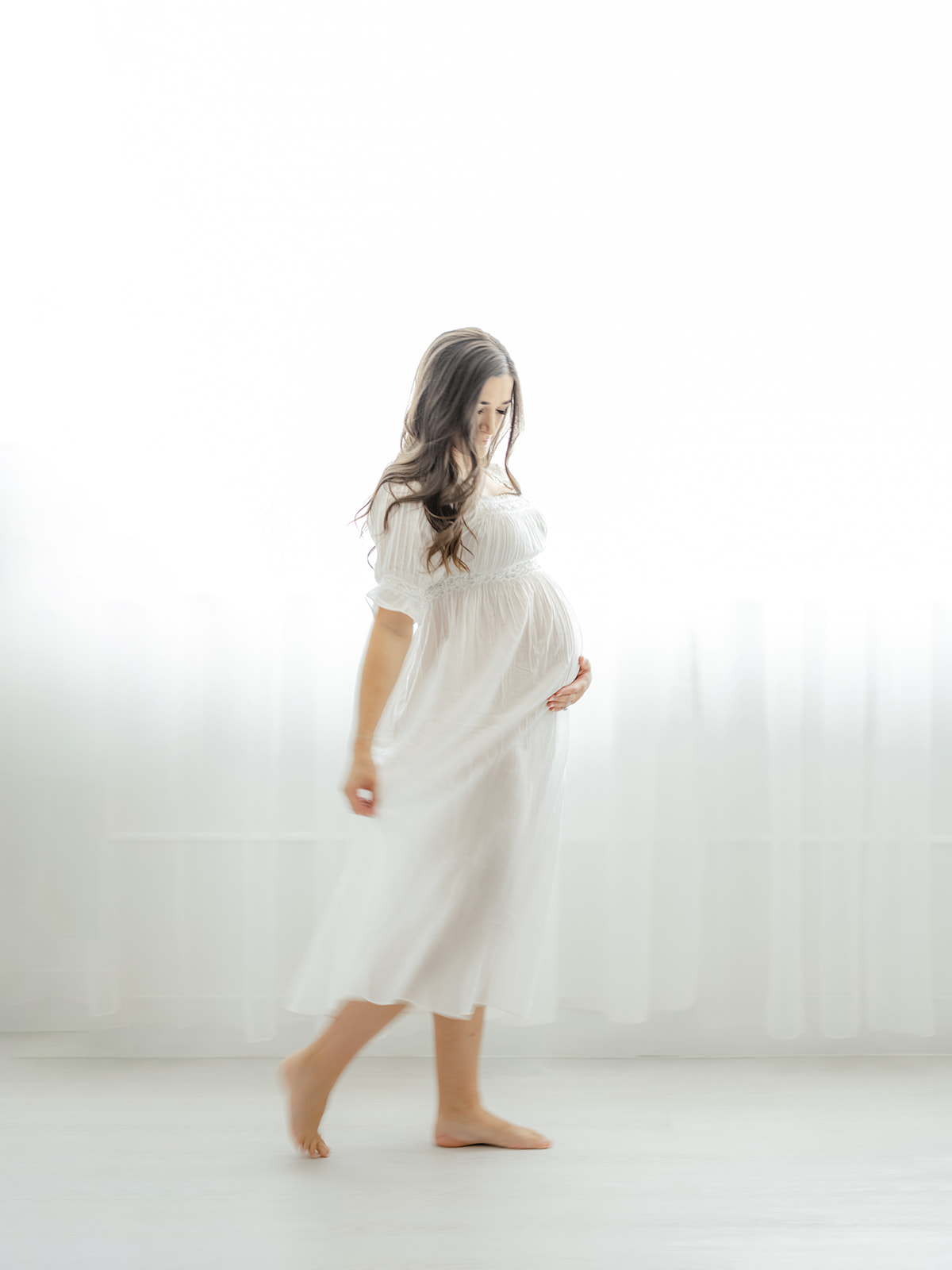 A mother to be walks in a studio in a white maternity dress after meeting Dallas Midwives