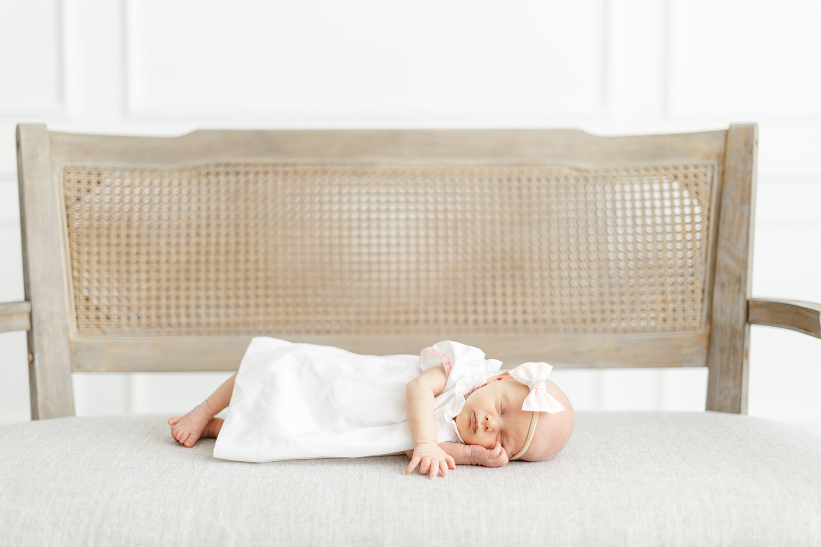 A newborn baby in a white dress and bow sleeps on a bench in a studio
