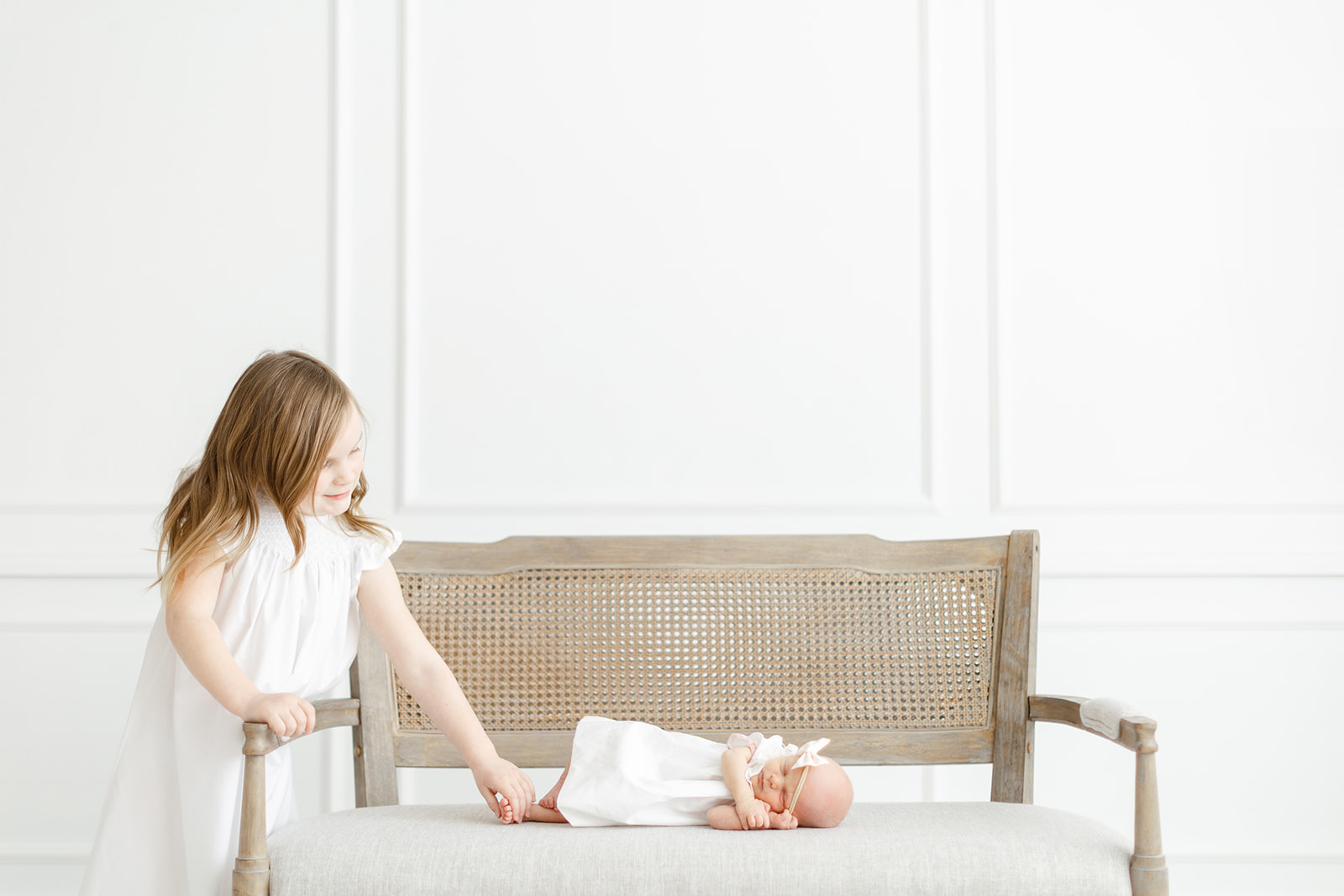 A newborn baby in a white dress lays on a bench in a studio while big sister holds her feet