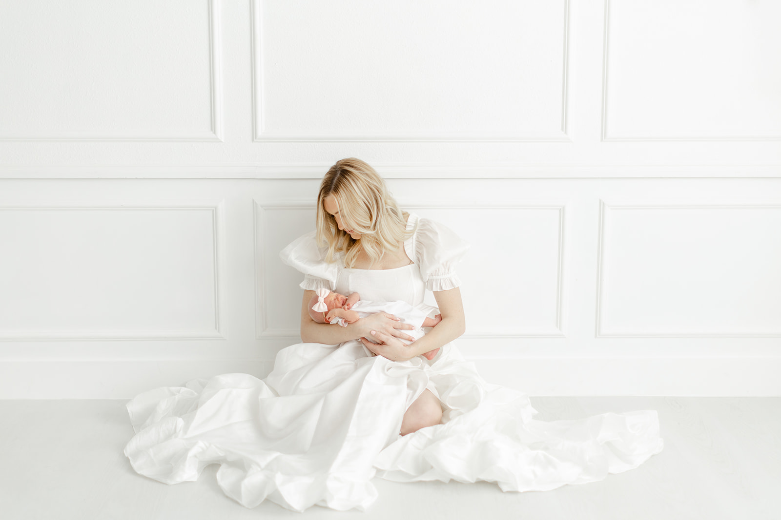 A mother in a long white dress sits on the floor of a studio cradling her newborn baby in her lap thanks to dallas lactation consultants
