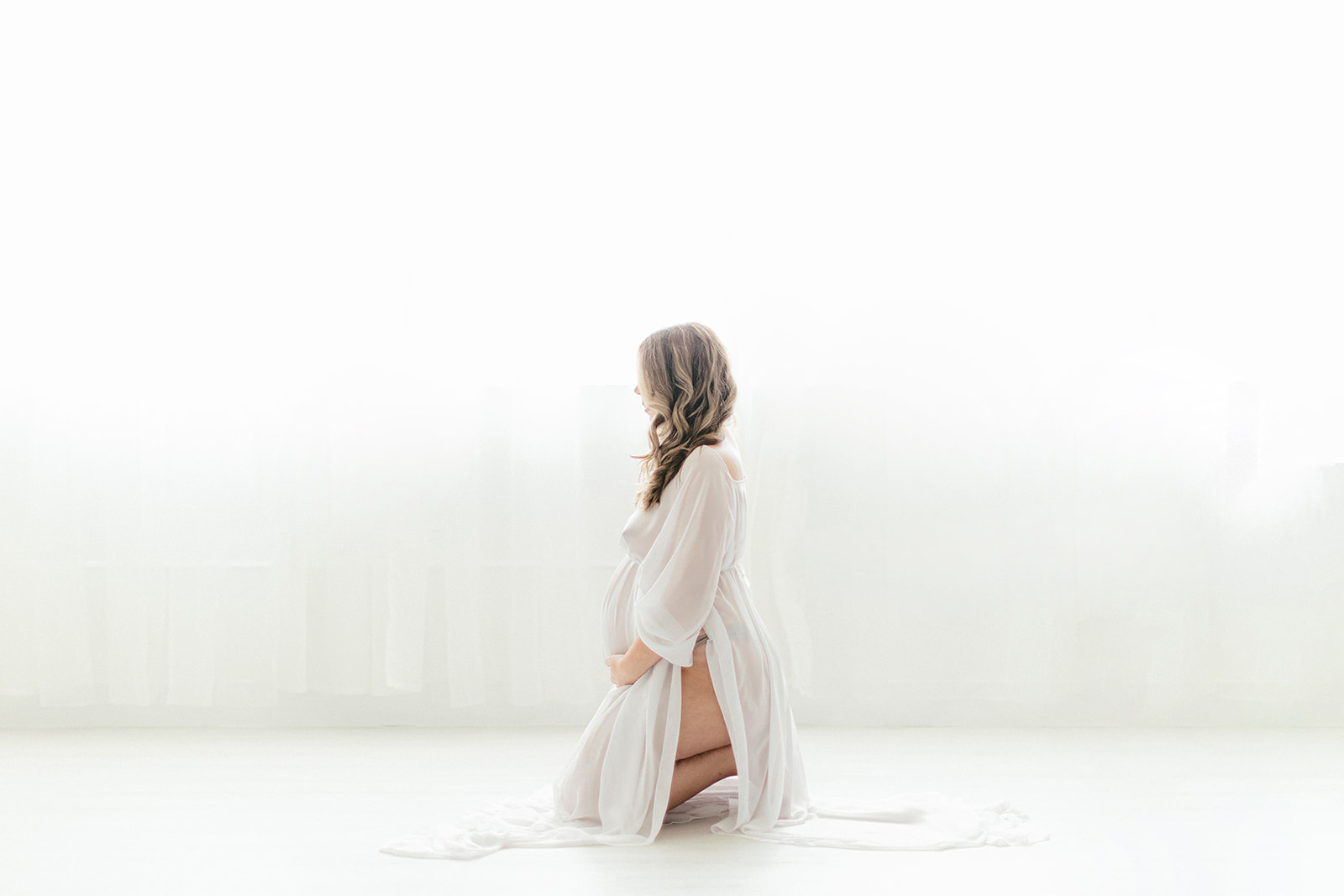 A mom to be in a sheer white maternity gown kneels on the floor of a studio holding her bump
