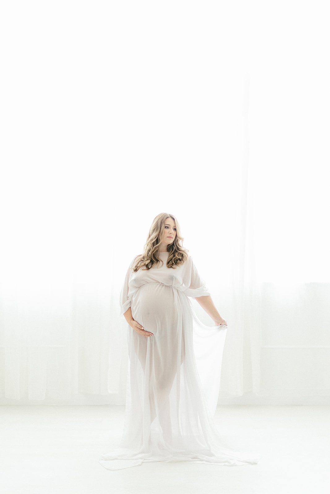 A mother to be in a white sheer maternity gown stands in a white studio holding her train before a 3D Ultrasound Dallas