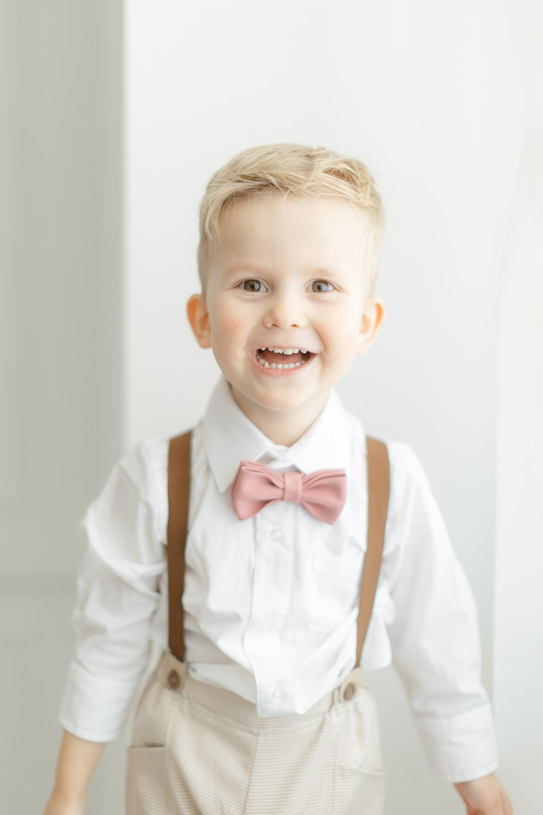 A happy toddler boy wearing a pink bowtie and suspenders stands in a studio smiling before visiting Dallas Daycares