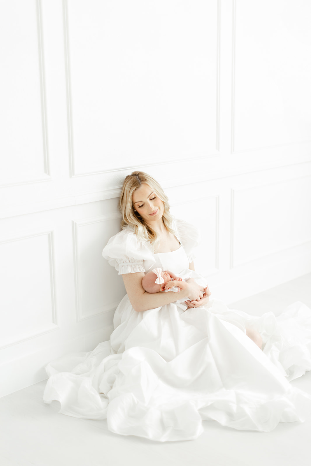 A happy mother in a white dress sits on the floor of a studio smiling down at her sleeping newborn baby in her arms thanks to Dallas Fertility Clinic