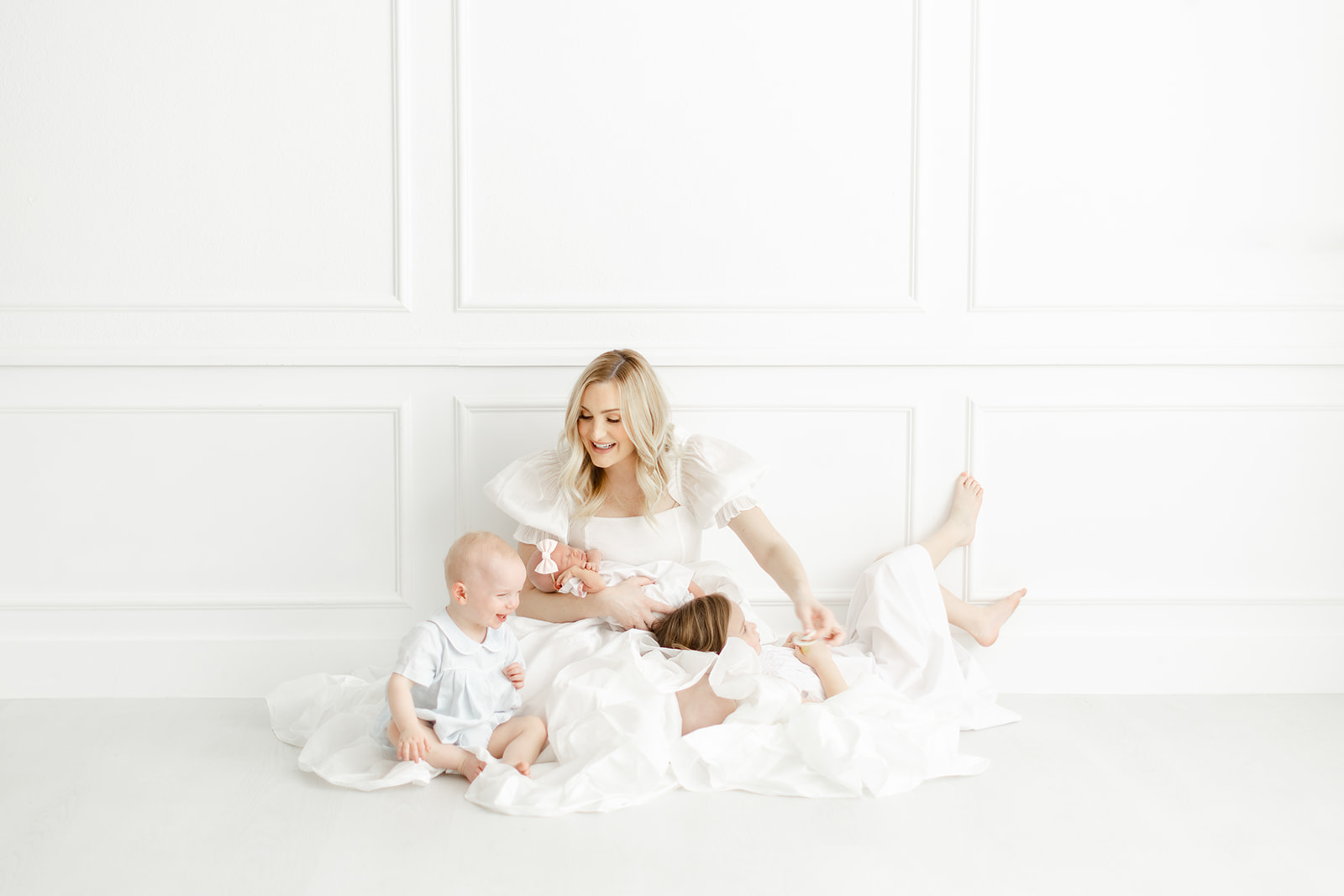 A mother in a white dress sits on the floor of a studio tickling and playing with her 2 toddlers and holding a sleeping newborn thanks to a Dallas Fertility Clinic