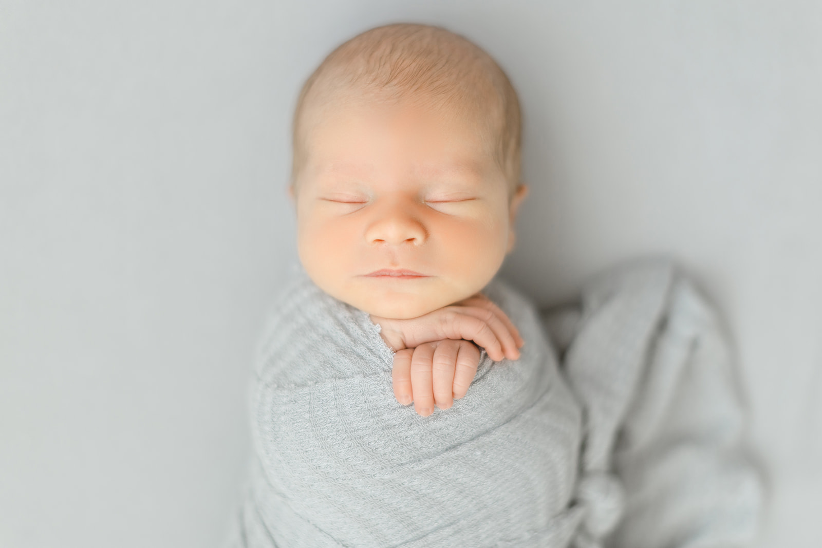 A newborn baby sleeps in a grey swaddle with hands sticking out in a studio