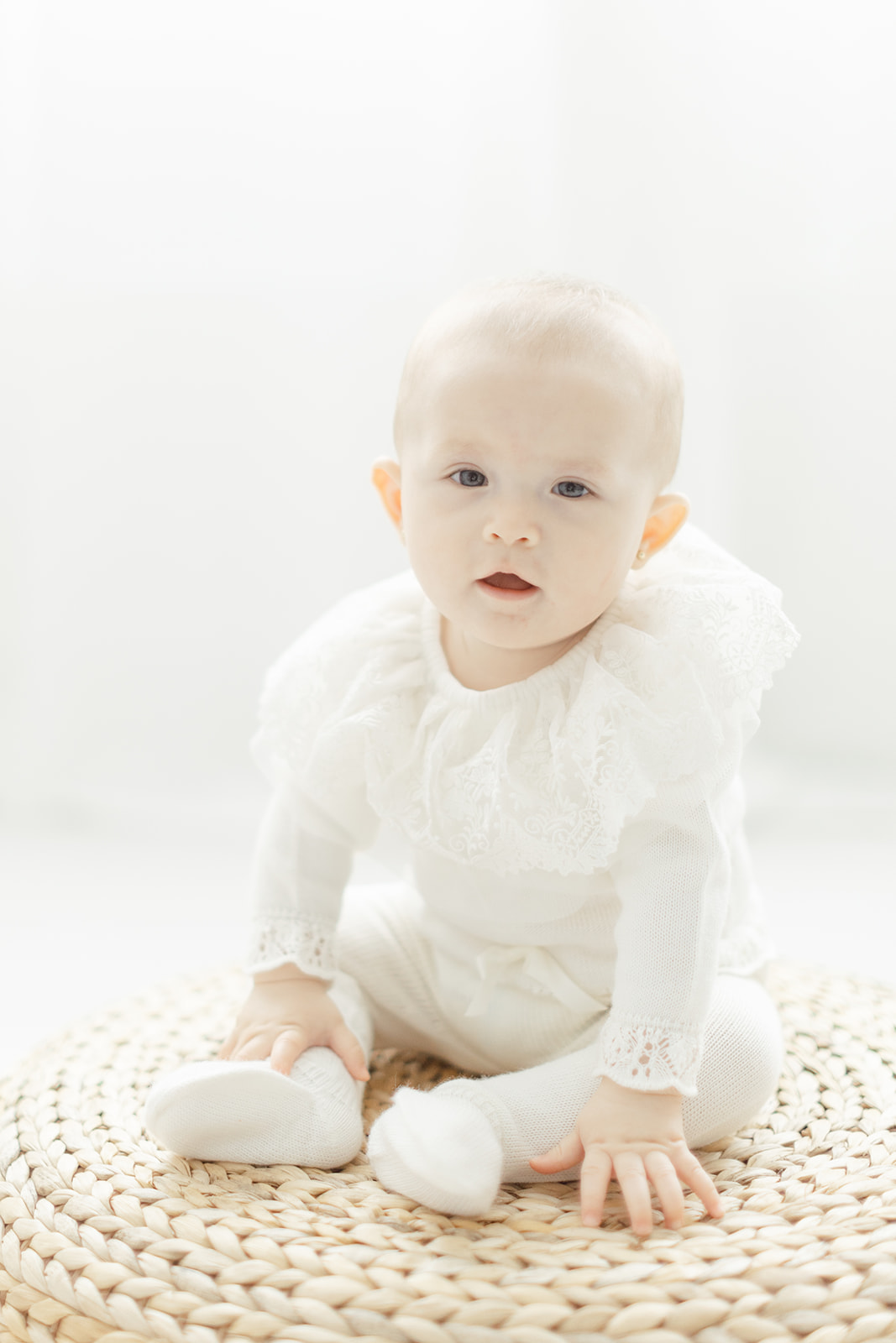 A toddler in a white onesie with a large collar sits on a woven stool under a window in a studio after visiting Dallas baby boutiques