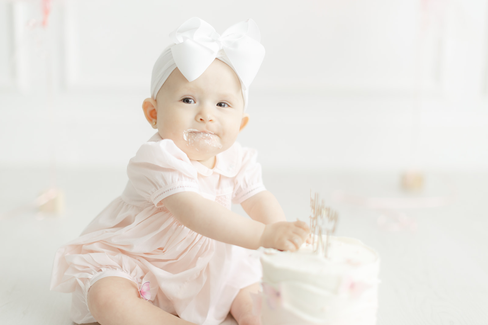 A toddler girl in a pink dress and large bow plays with a cake in a studio after visiting Dallas baby boutiques