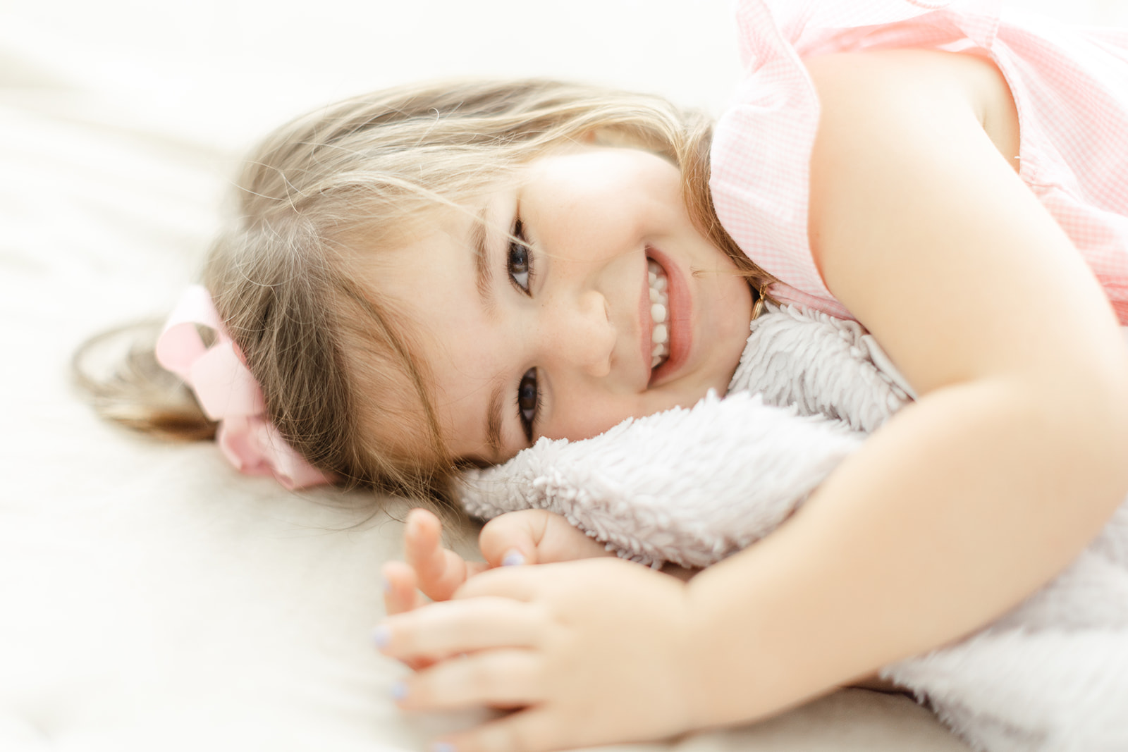 A toddler girl in a pink dress smiles big while playing on the floor with a blanket