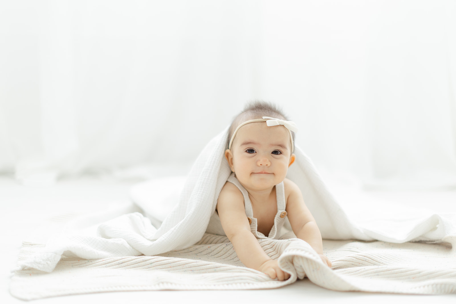 An infant girl in white overalls and bow plays under a blanket on the floor of a studio after meeting Dallas Pediatricians