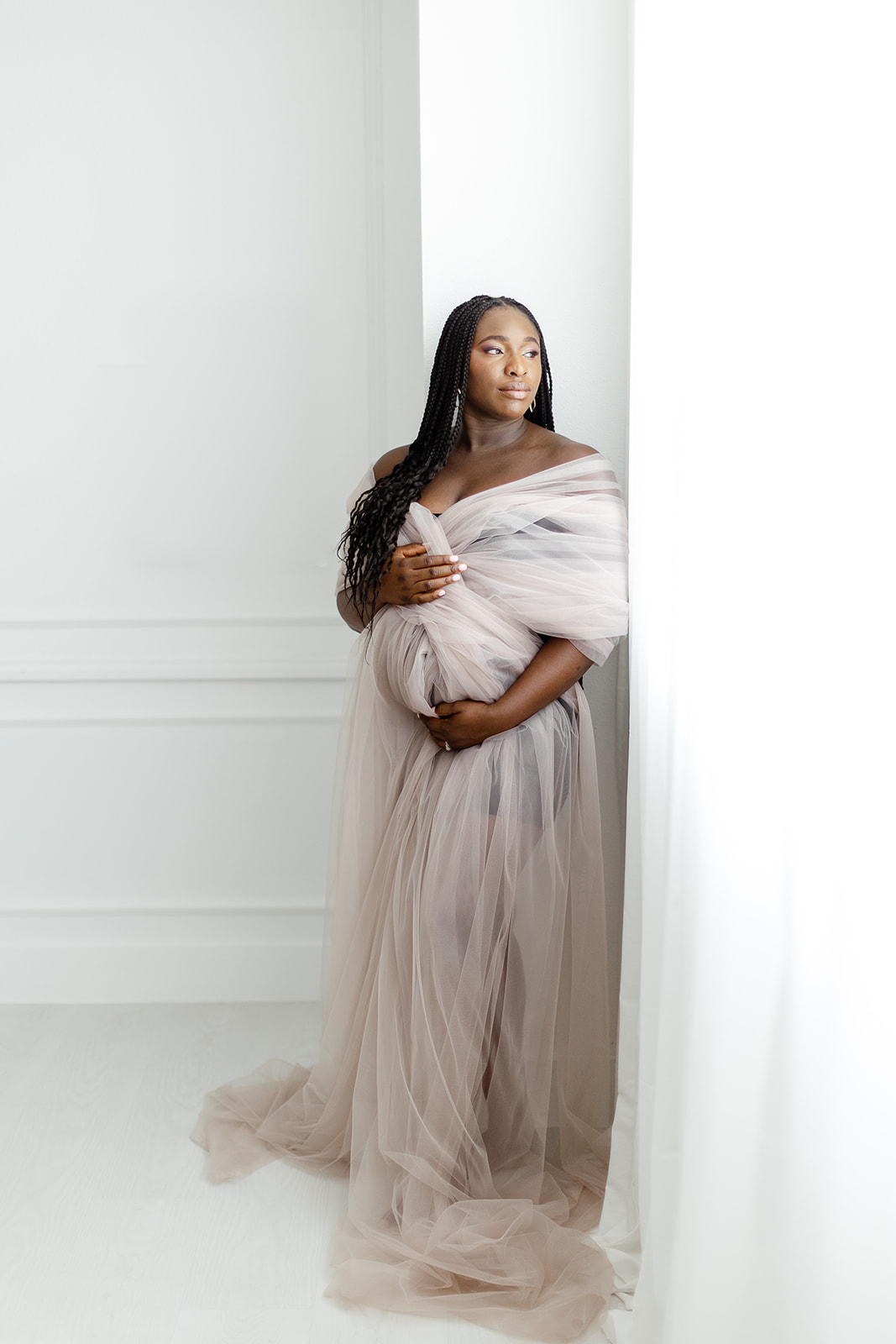 A mother to be in a pink tule maternity gown smiles out a window in a studio before a Dallas prenatal massage