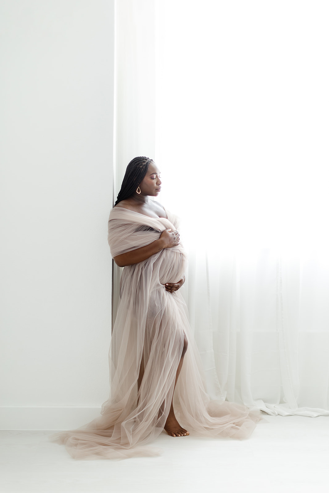 A mom to be in a pink maternity gown stands in a window of a studio holding her bump before a Dallas prenatal massage