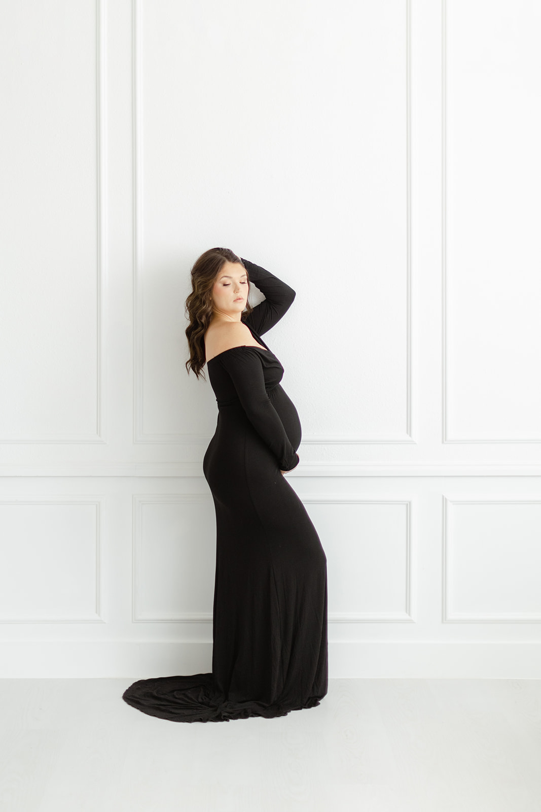 A glamorous mom to be stands in a studio holding back her hair and a hand under the bump