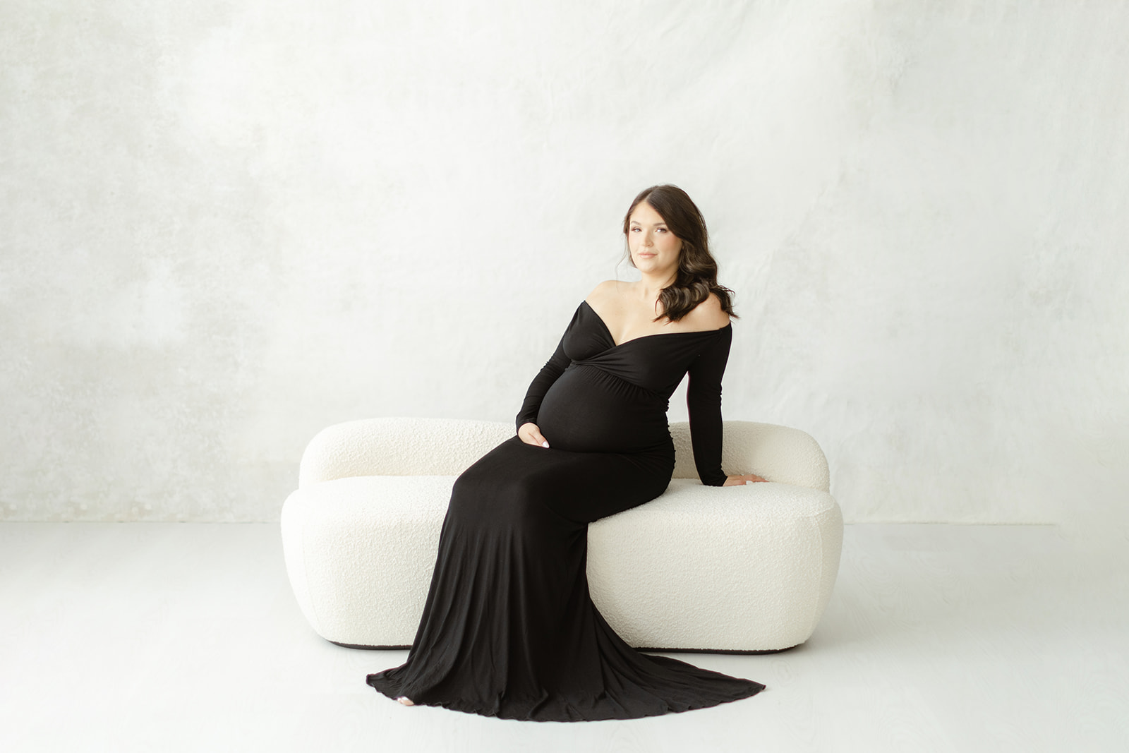 A happy mother to be in a long black maternity gown sits on a white bench in a studio holding her bump thanks to Fertility Acupuncture Dallas