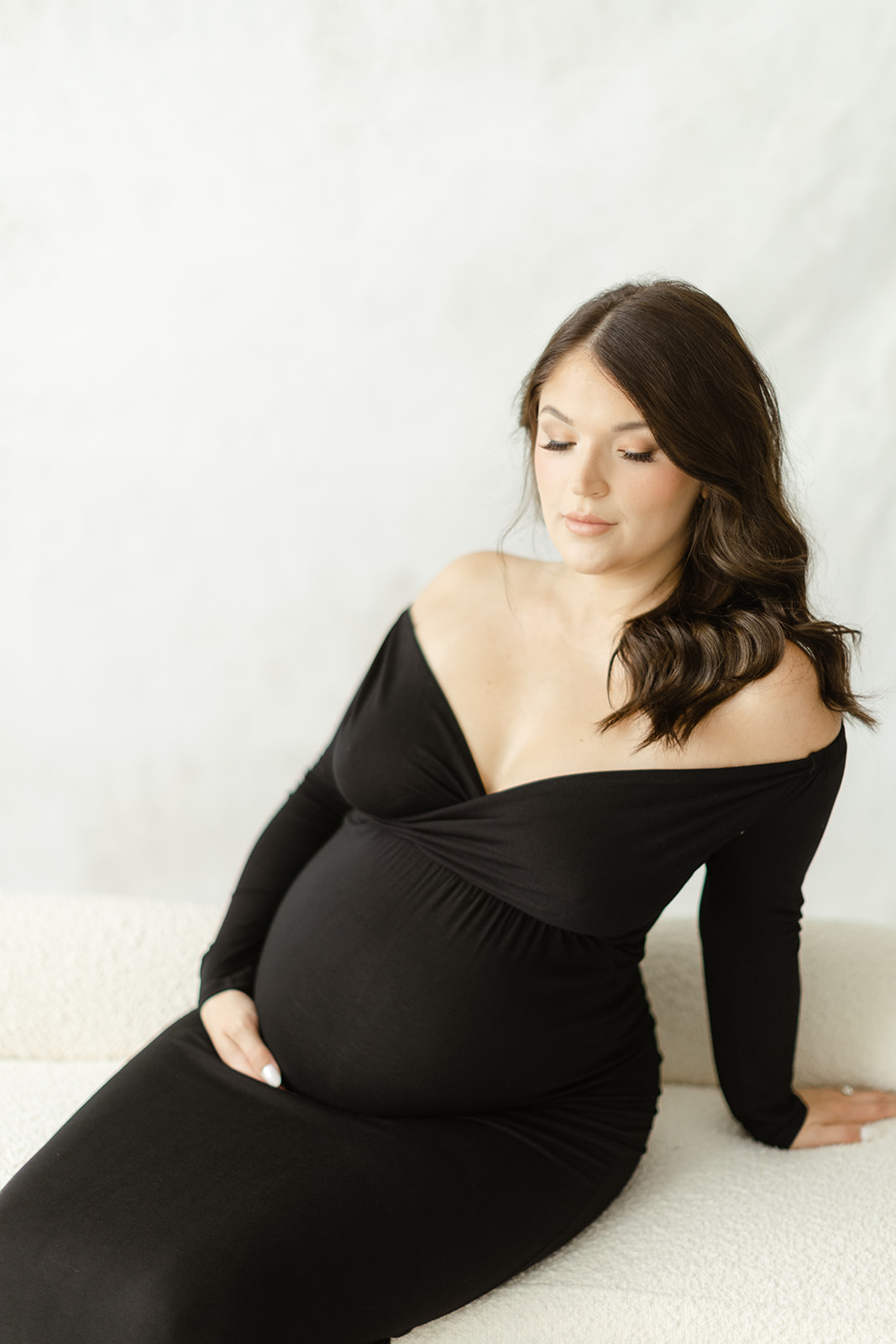A mom to be in a black maternity gown sits on a bench in a studio leaning back on one arm thanks to Fertility Acupuncture Dallas