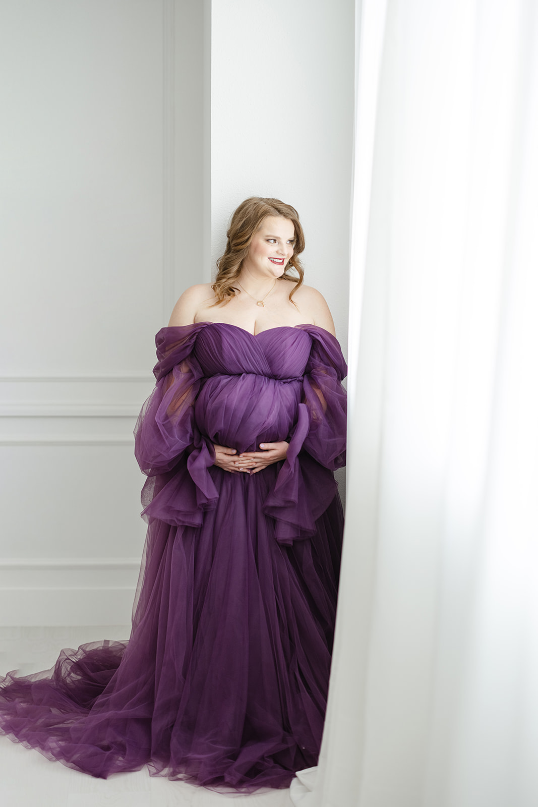 A mom to be leans against a wall smiling out a window while holding her bump in a long tule maternity gown