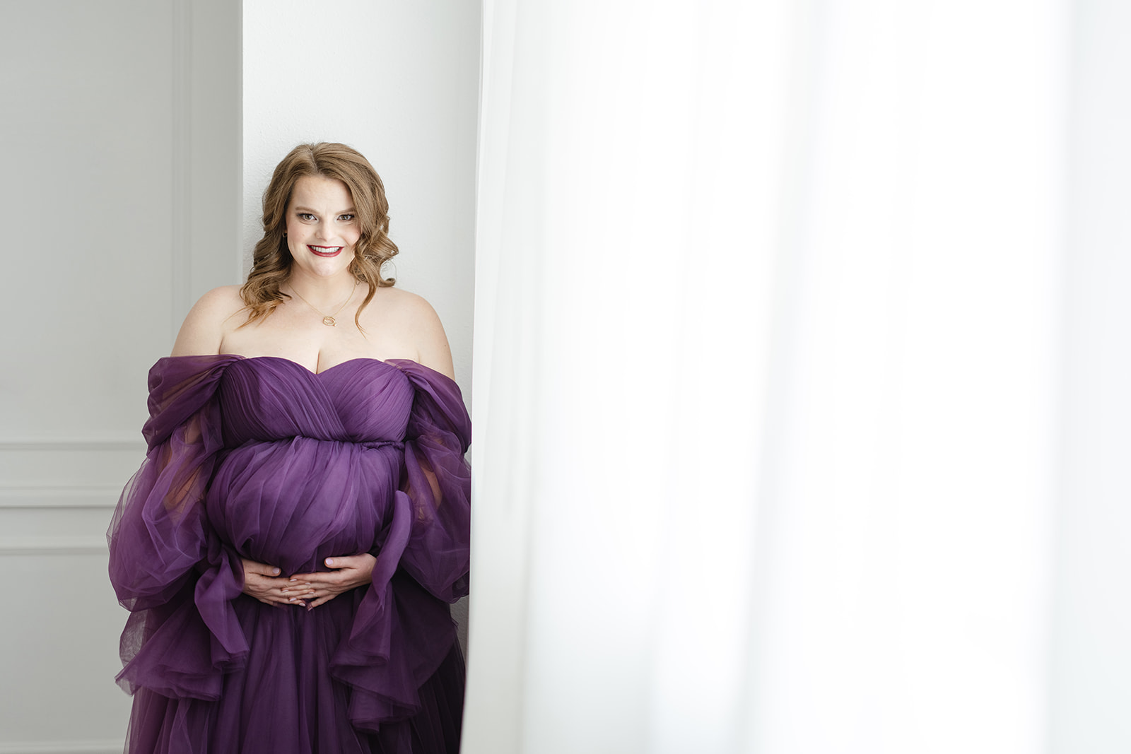 A happy expecting mother stands in a studio holding her bump in a purple tule maternity gown before visiting a Prenatal Chiropractor Dallas