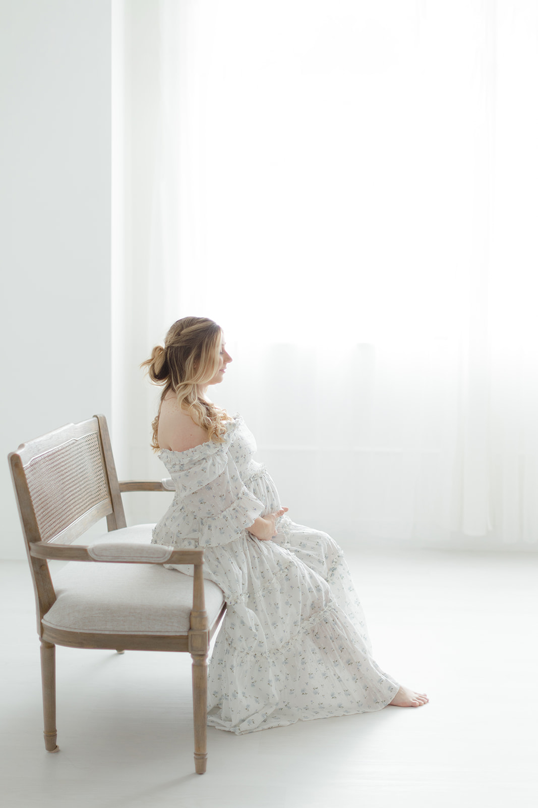 A pregnant woman sits on a bench in a studio in a white maternity dress holding her bump
